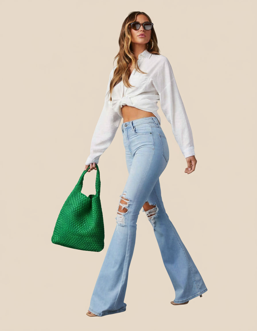 Woman wearing a white blouse and light blue jeans, holding a green handbag against a tan background