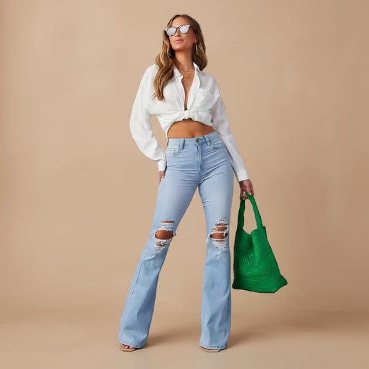 Woman wearing a white blouse and light blue jeans, holding a green bag against a tan background